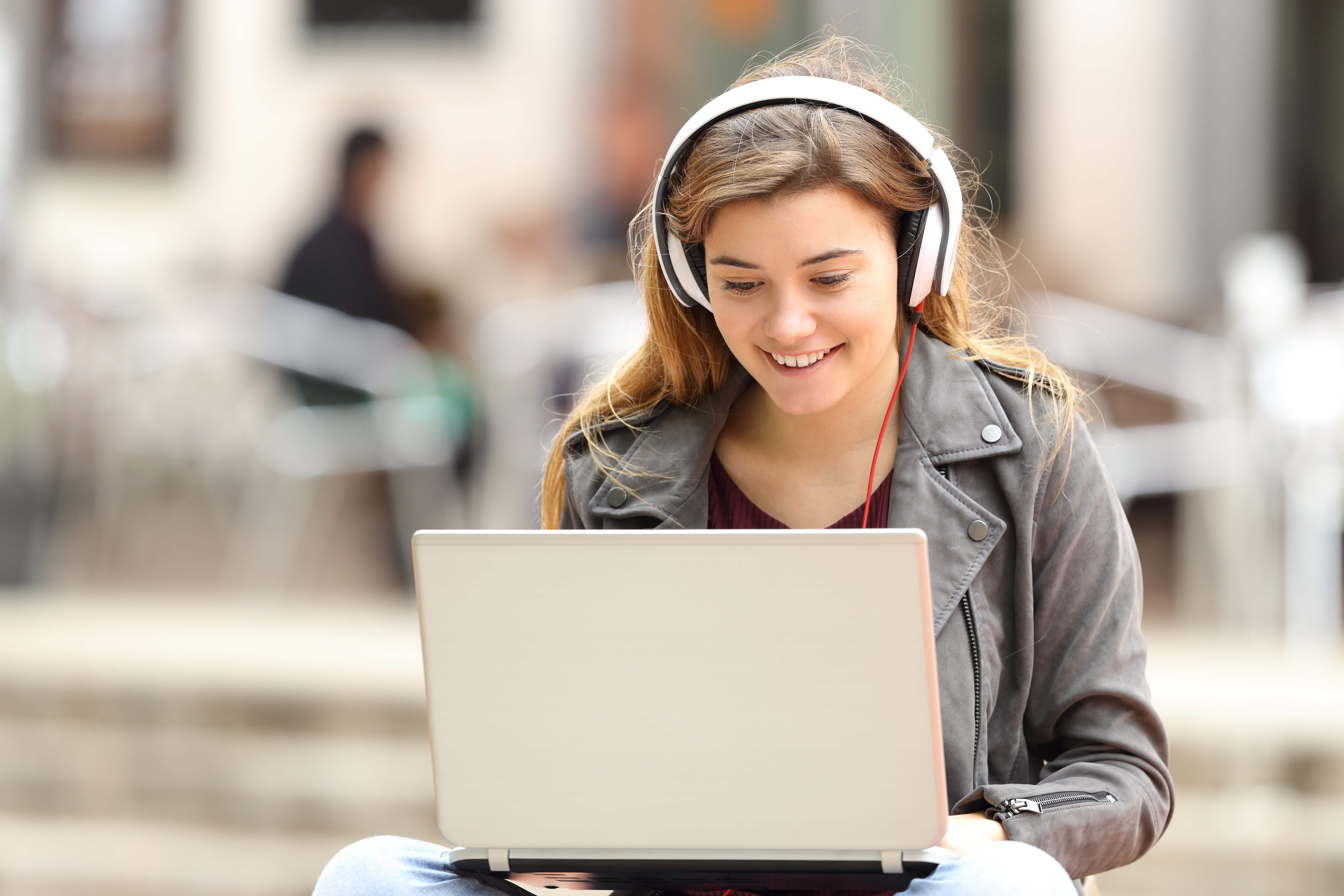 Casual girl listening music with headphones and searching songs in a laptop sitting on a bench in the street