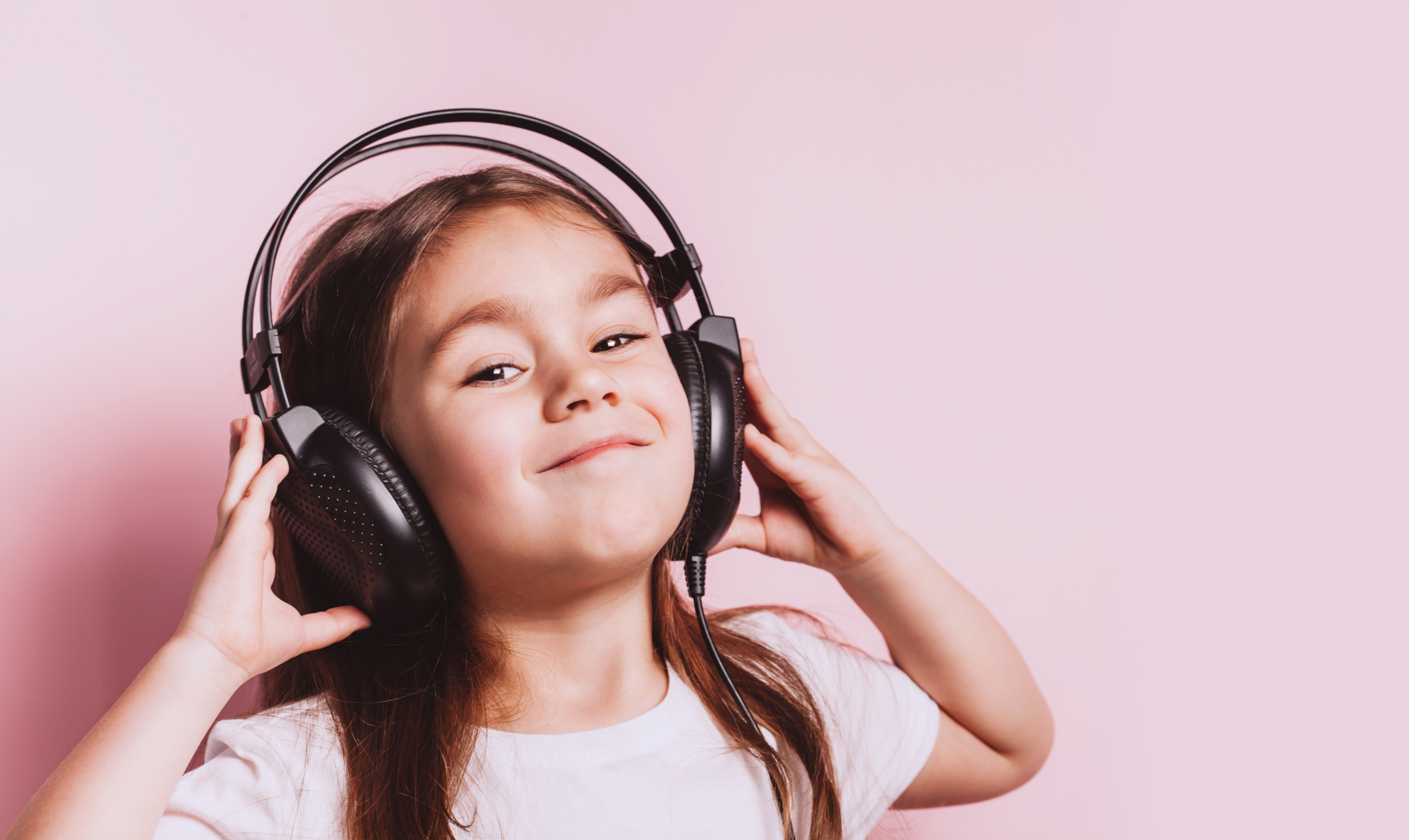 Cute little girl listening music wearing earphones on pink background. Funny emotions. Copyspace for text.